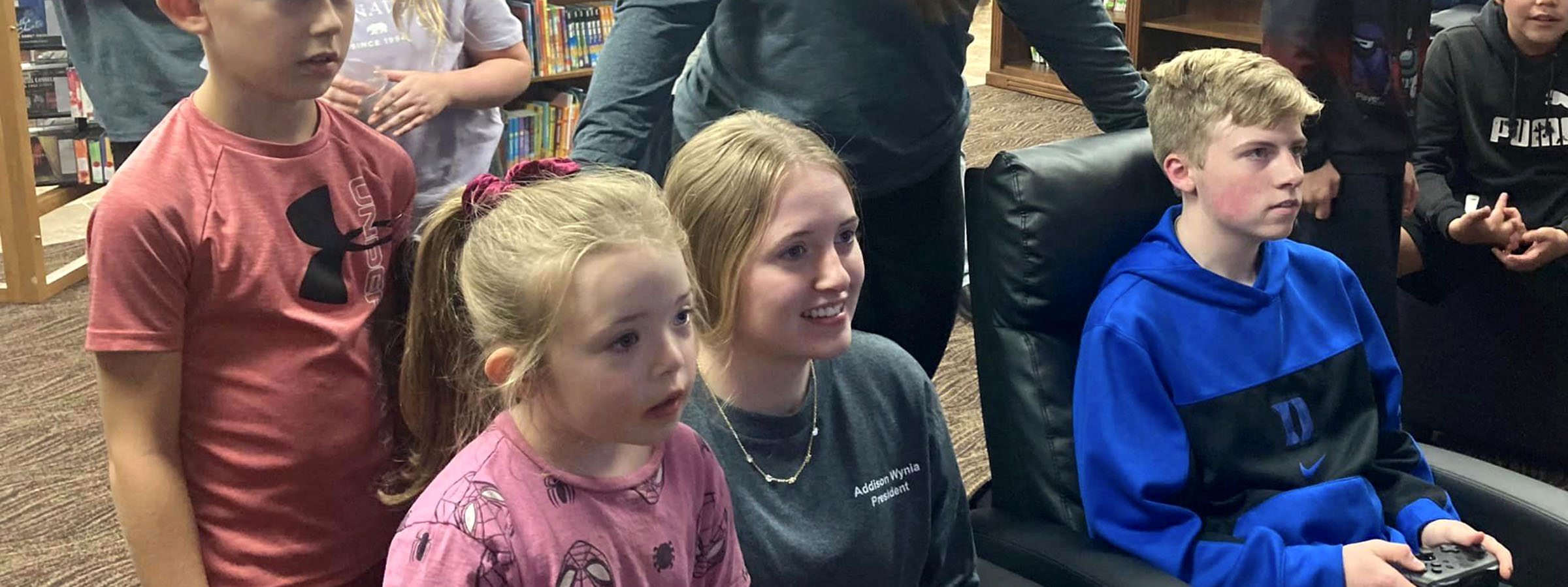 A group of children gathered in a library, listening attentively to a man seated in a chair, surrounded by books.