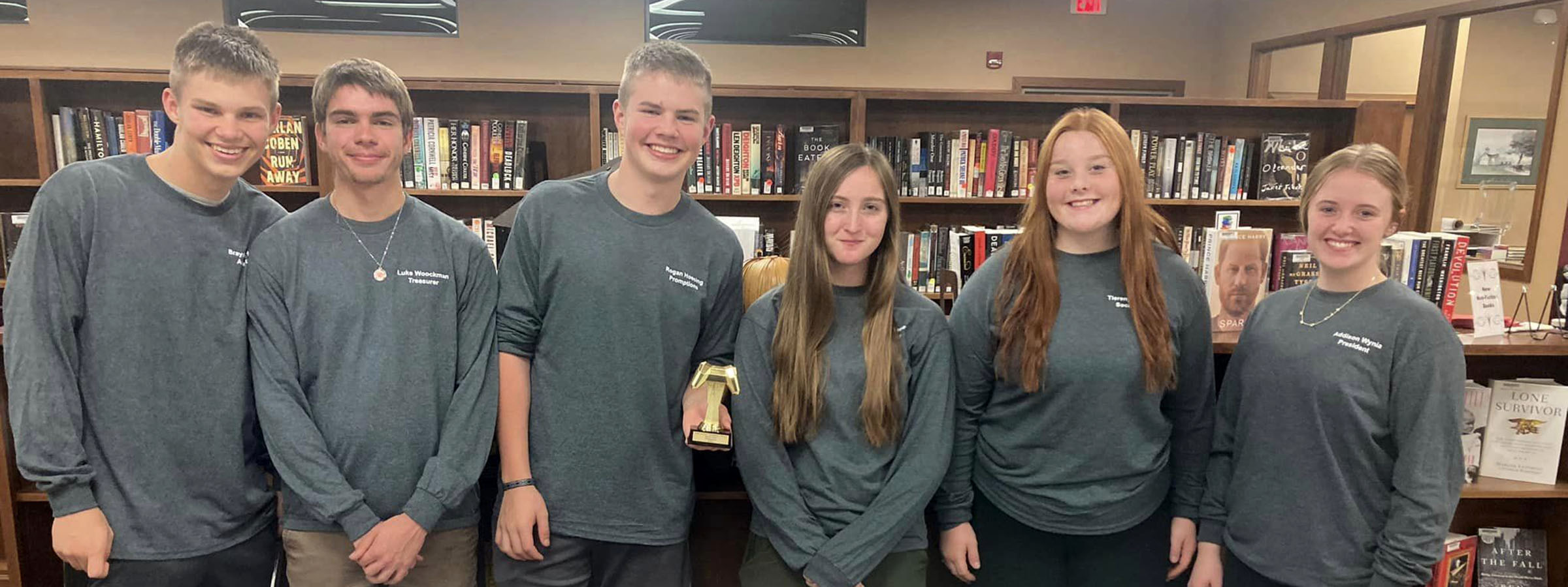 Five young people in gray shirts standing in front of bookshelves.