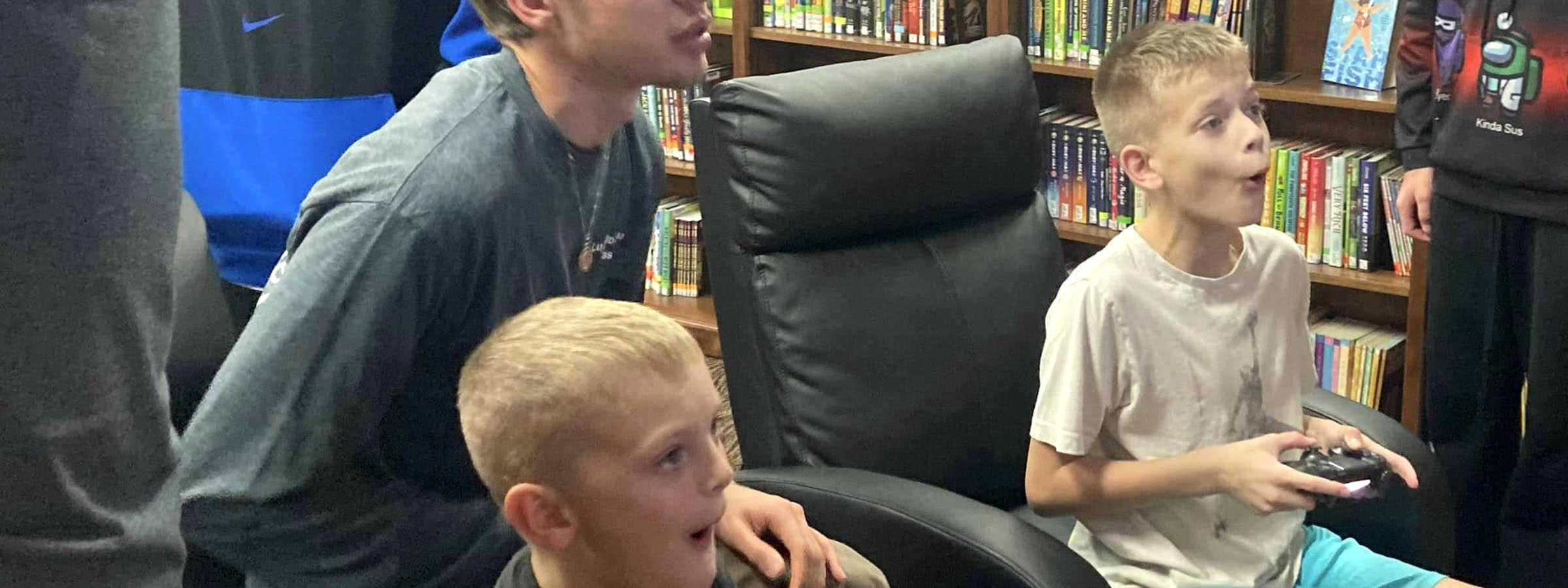 Boys playing video games in library, surrounded by books and computers.