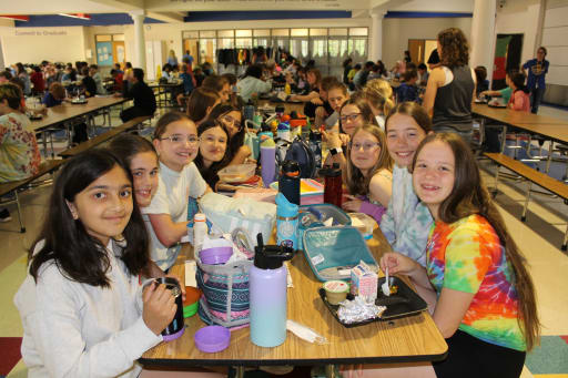 A group of smiling girls sitting together at a lunch table in a school cafeteria, with various lunch items and water bottles in front of them.
