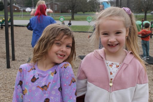 Two young girls smile at the camera while standing on a playground, with other children playing in the background.