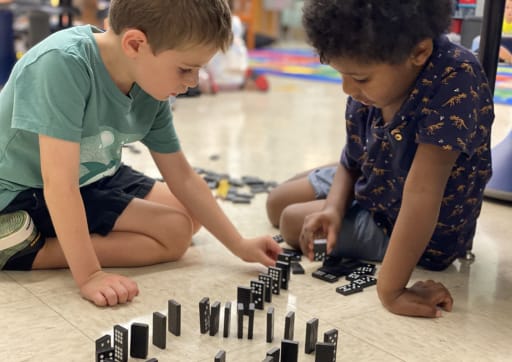 Two children are sitting on the floor, focused on arranging black dominoes in a line, with a colorful classroom background.