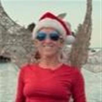 A woman wearing a Santa hat, sunglasses, and a red shirt, smiling outdoors near a tree.