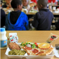 This image shows another nutritious school lunch being served in a cafeteria setting, with students visible in the background.This meal includes:  Grain & protein (pizza)  Vegetables (salad + peas)  Fruit (orange slices)  Dairy (milk)