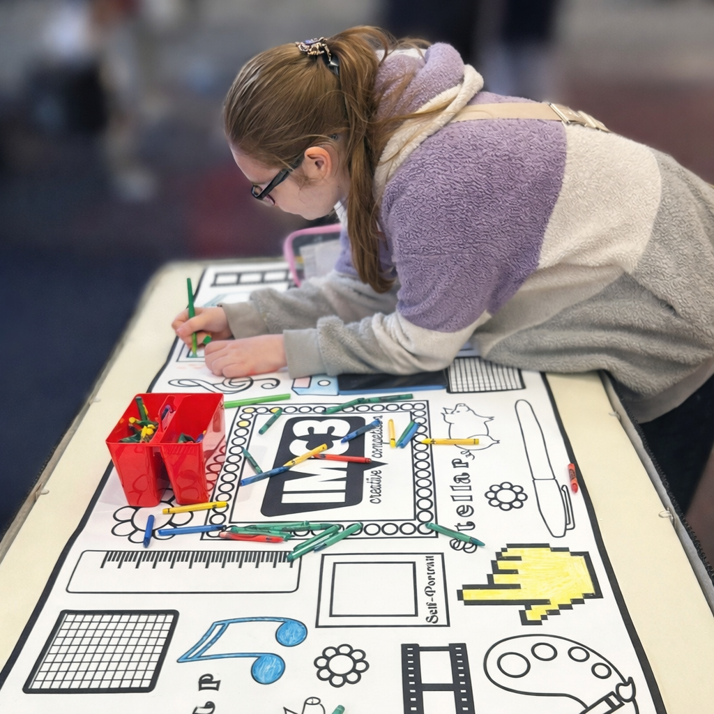 Student coloring a large arts-themed poster at a table, with crayons spread out, while the background is softly blurred.