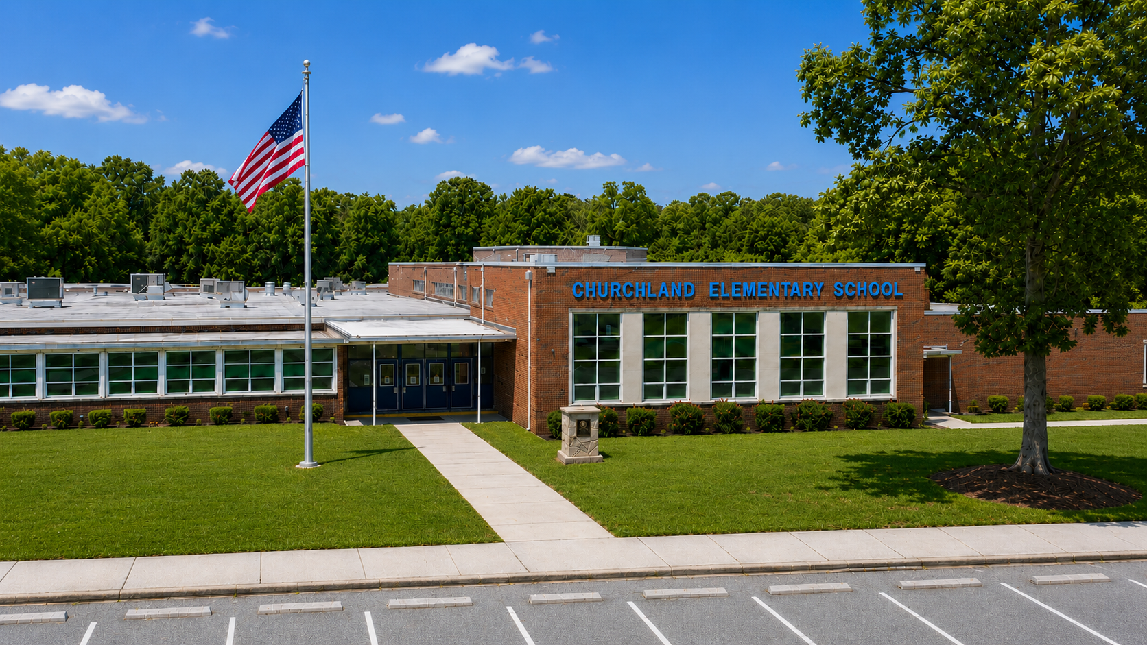 A school building with a flag and large windows, surrounded by grass and trees, and a parking lot.