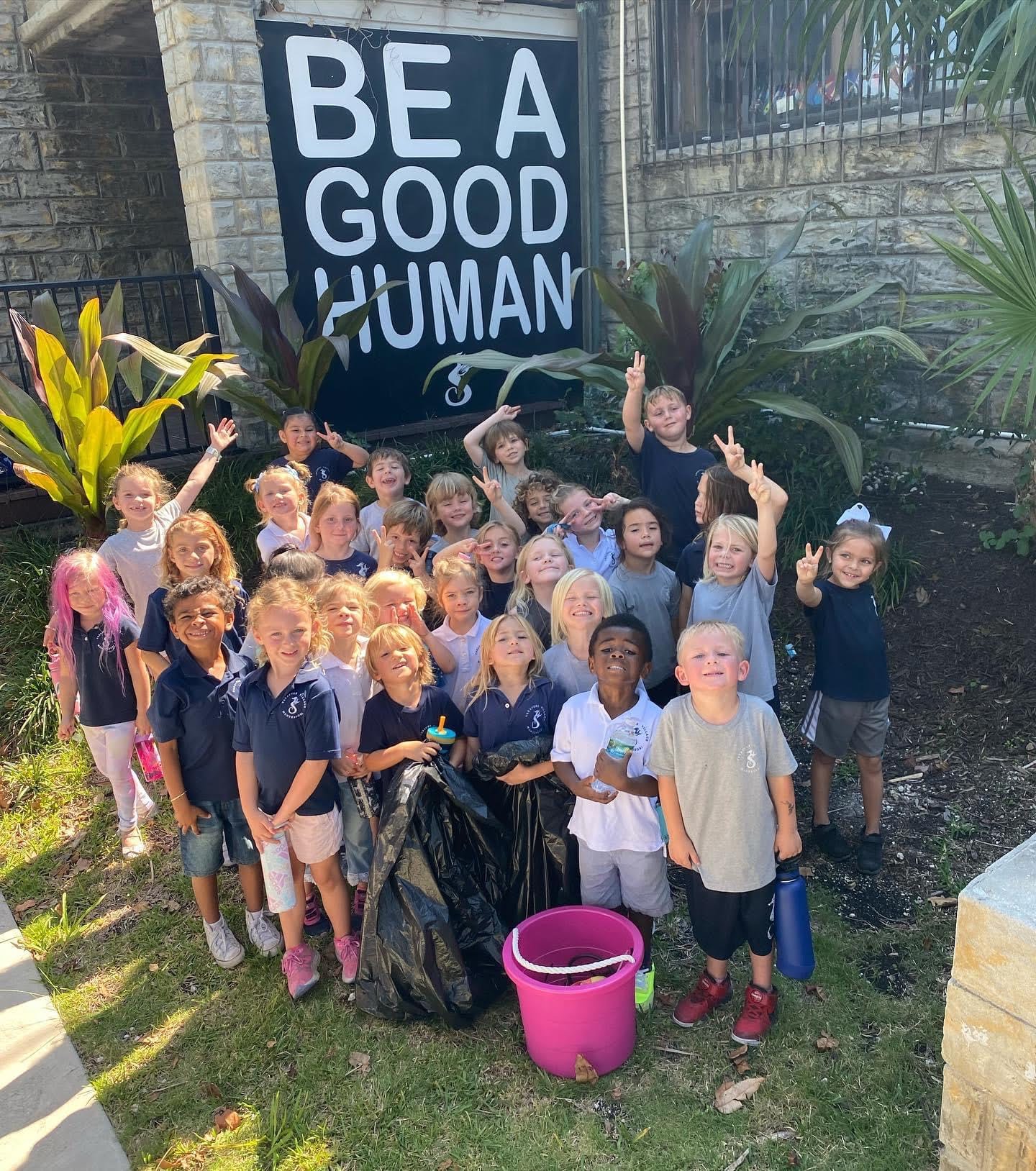 A group of children smiling and posing for a photo in front of a large sign that reads 'BE A GOOD HUMAN', with some holding trash bags and a pink bucket.