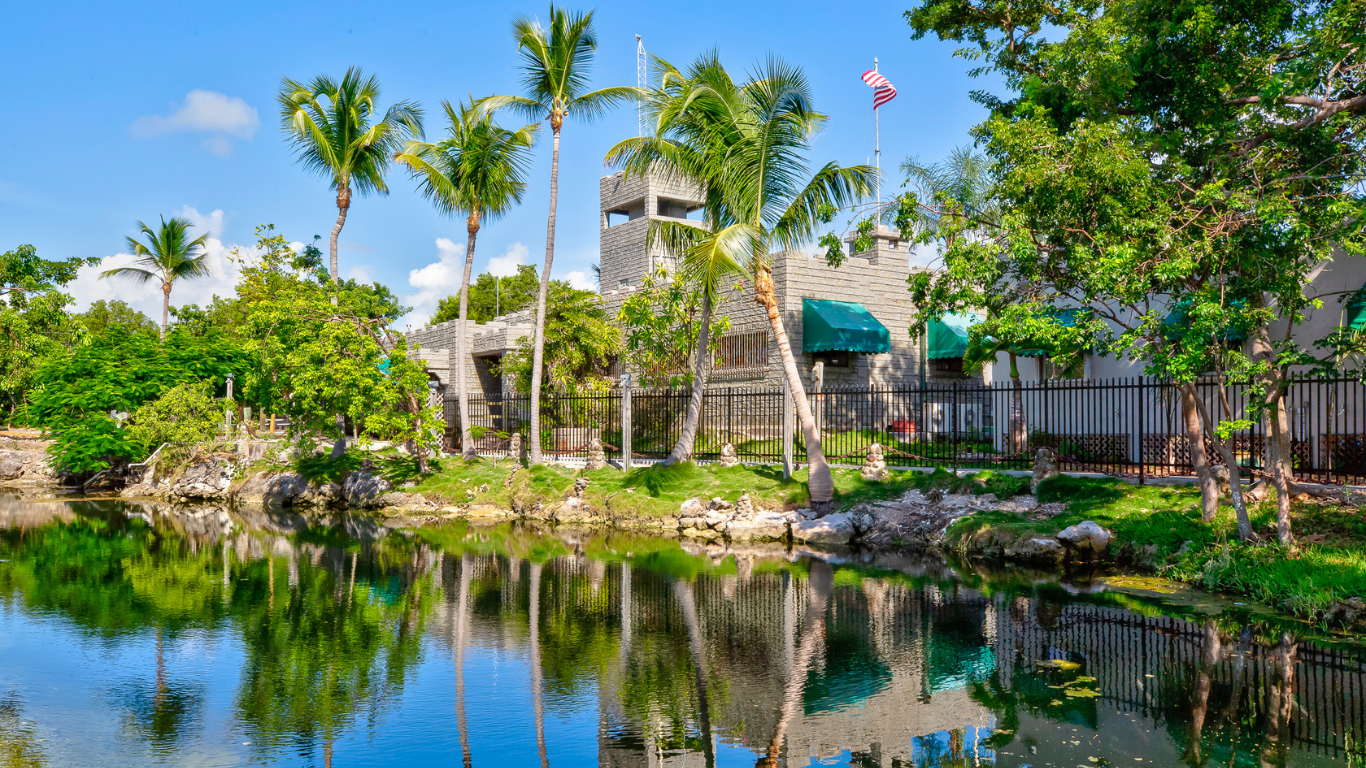 A serene landscape featuring a building surrounded by palm trees and lush greenery, reflecting in a calm body of water under a clear blue sky.
