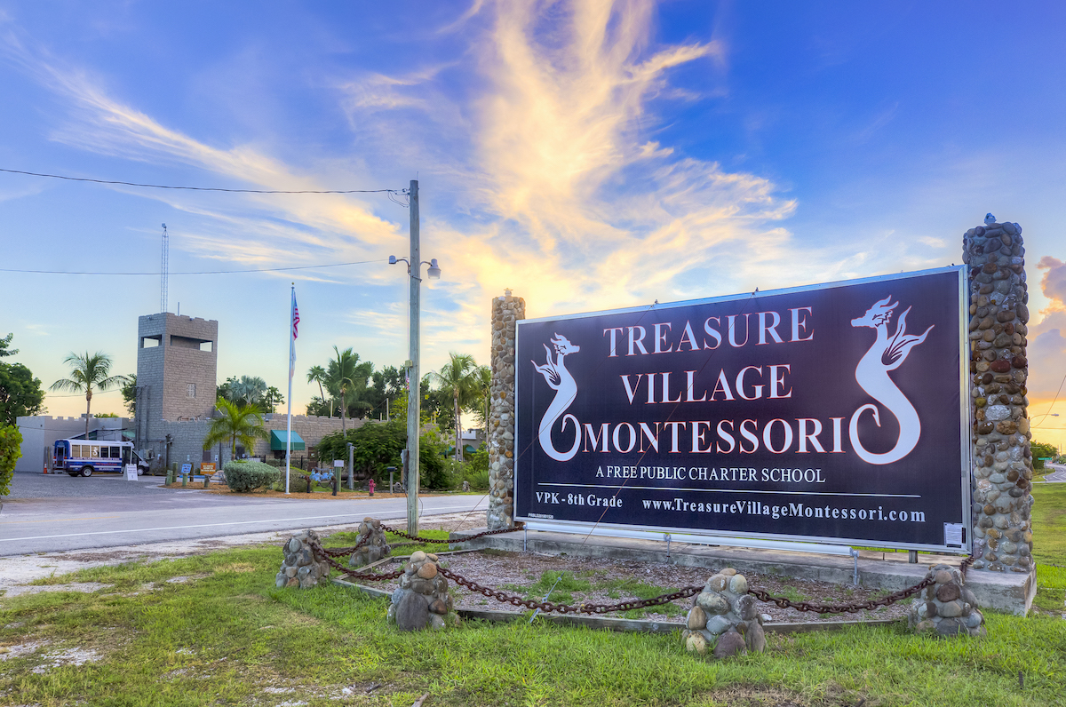 A large sign for Treasure Village Montessori, a public charter school, is displayed against a colorful sky with clouds, alongside a building and palm trees in the background.