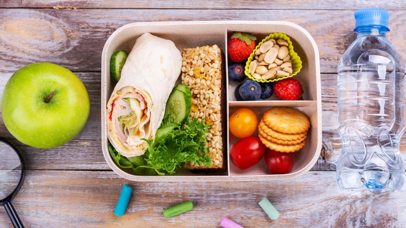 A lunchbox filled with a wrap, a grain bar, assorted fruits, cookies, and a bottle of water, accompanied by a green apple and colorful chalk pieces on a wooden surface.