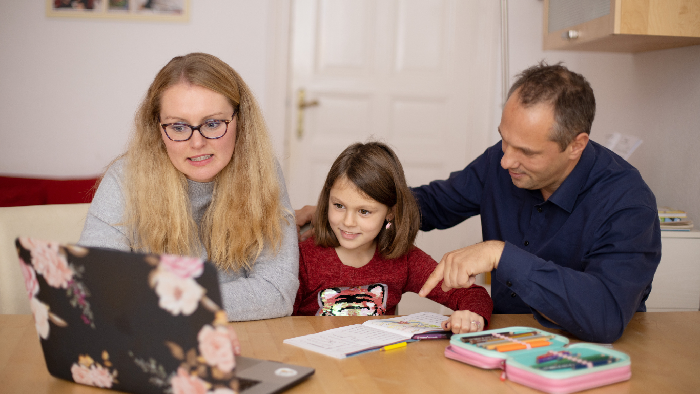 A woman and a man assist a young girl with her homework at a table, while a laptop displays educational content in front of them.