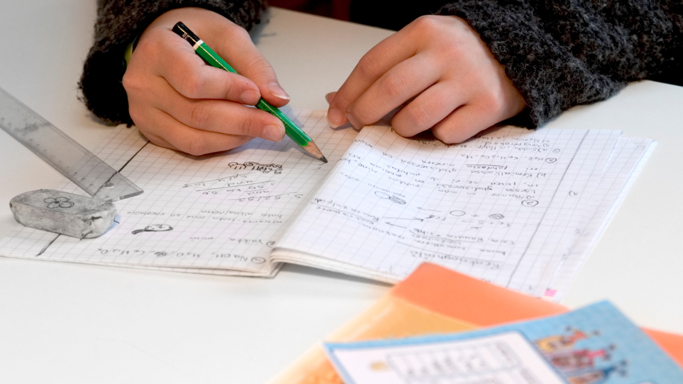 A person is writing in a notebook with a green pen, surrounded by various colored papers and a ruler on a white table.