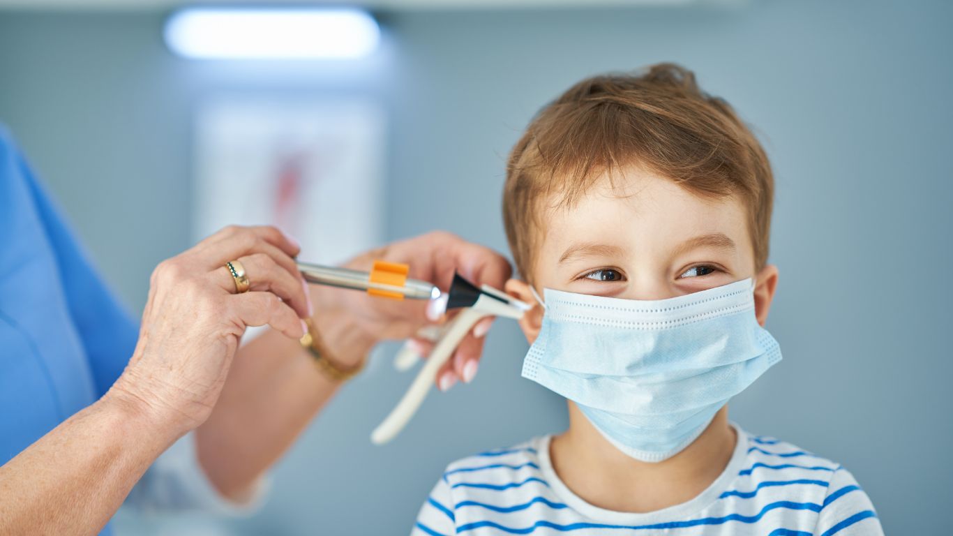 A young boy wearing a blue mask smiles while a healthcare professional prepares to administer a vaccine using a syringe near his ear.