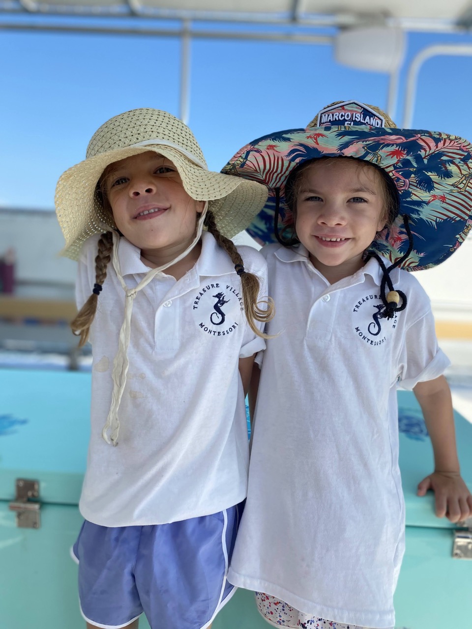 Two young girls wearing matching white polo shirts and large sun hats, smiling and standing close together on a boat deck under a clear blue sky.