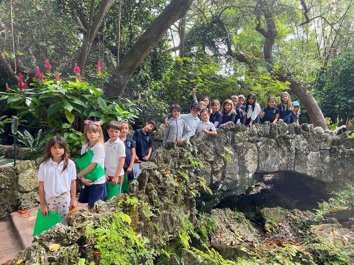 A group of children stands on a stone bridge surrounded by lush greenery and tropical plants in a garden setting.