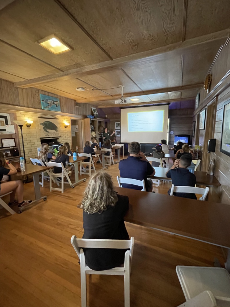 A group of people seated in a room with wooden walls, attending a presentation projected on a screen at the front.