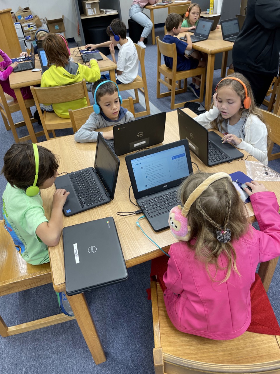 A group of children wearing headphones are focused on their laptops at a table in a classroom setting.
