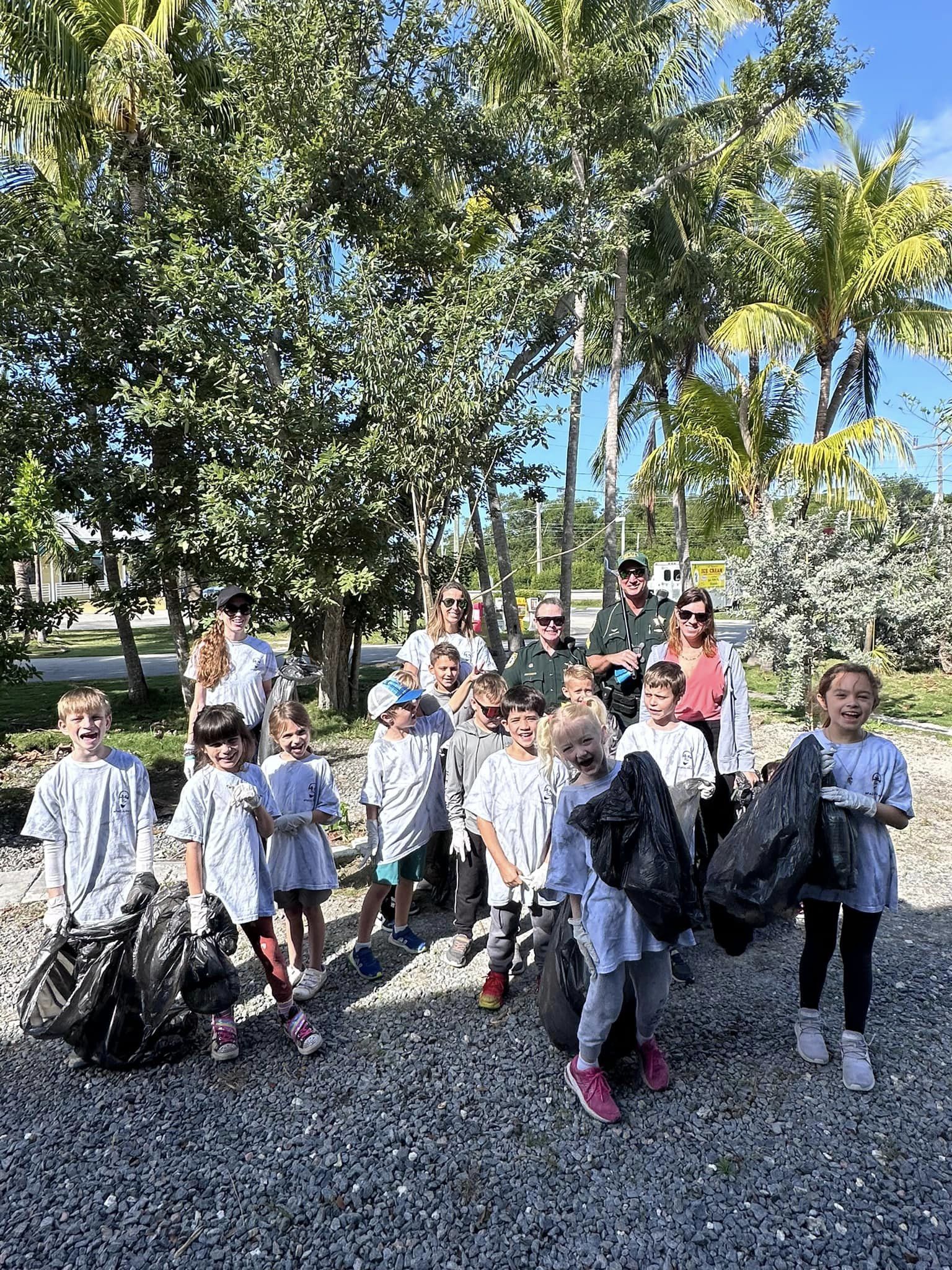 A group of children and adults, wearing matching shirts, stand together outdoors in a park-like setting with palm trees in the background, some holding bags, likely after a cleanup activity.