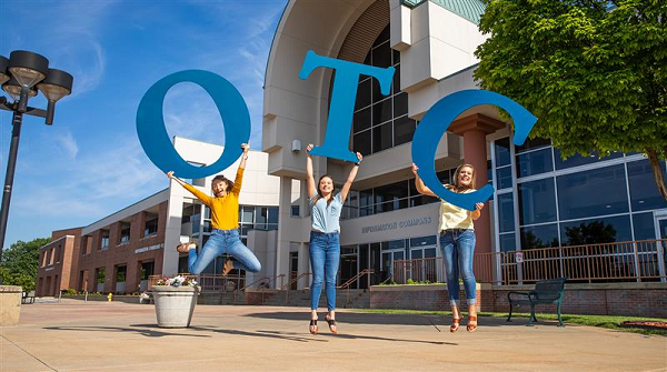 Three women joyfully jump in the air, forming the letters "OTC" with their bodies, celebrating together.