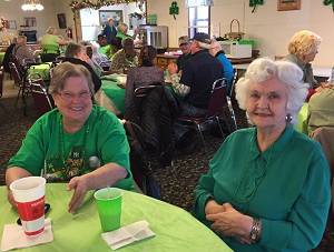 Two older women enjoying a conversation at tables adorned with vibrant green tablecloths in a cozy setting.