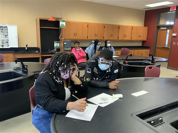 Two people wearing masks and engaged in a task at a table in a laboratory or classroom setting.