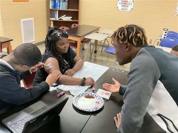 Three individuals in a classroom setting, focusing on an object with a syringe on a table.