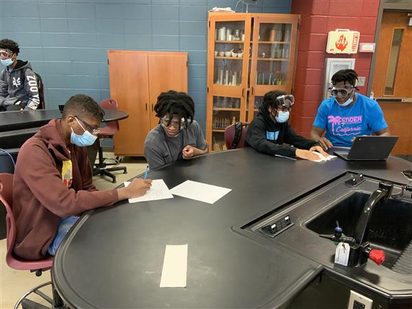 Group of students wearing masks and working on papers at a table in a classroom setting.