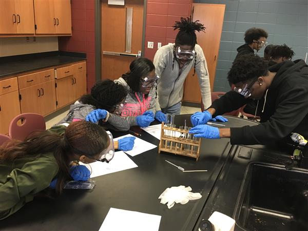 A group of students in a science lab, wearing safety goggles and gloves, working on projects together.