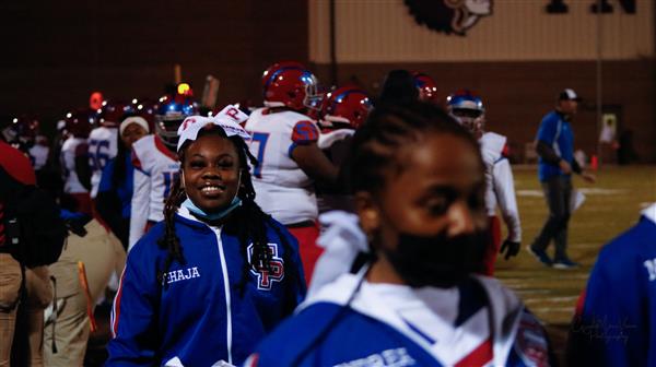 A young woman standing on a football field, wearing a blue and white jacket, smiling towards the camera.