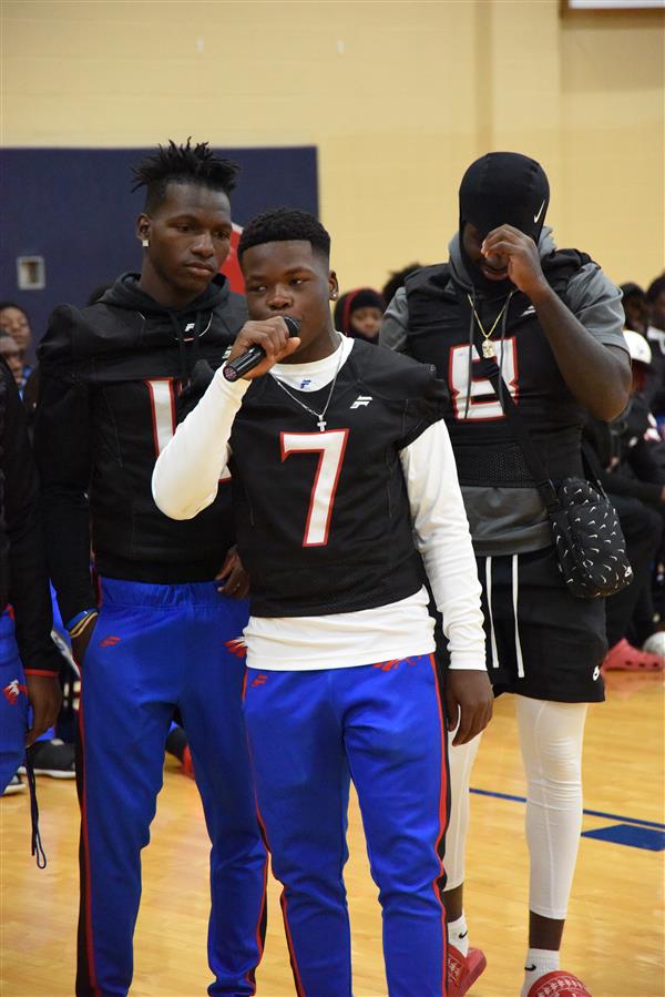 Young athletes in football uniforms, one delivering a speech while another watches with interest.