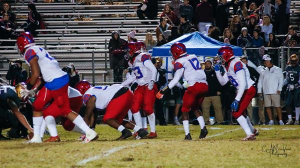 Action shot of a football game, with players in the middle of a play and spectators watching from stands.