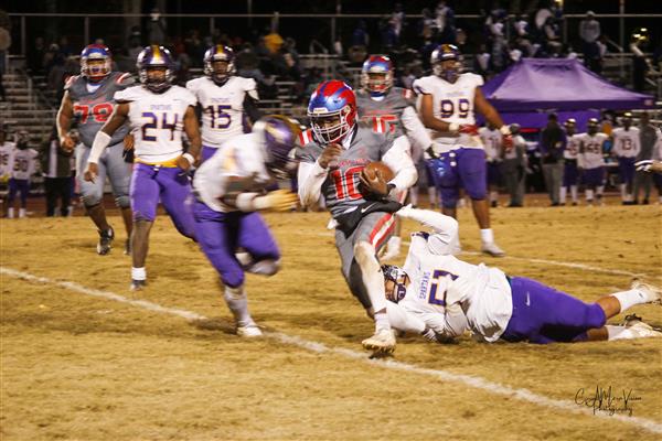 This is a photo of a high school football game in progress. The central figure, presumably the quarterback, is running towards the right with a football in hand while players in various positions are on the field around him.