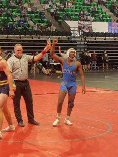 A male wrestler in a blue outfit celebrates with a man in a suit after winning a competition.