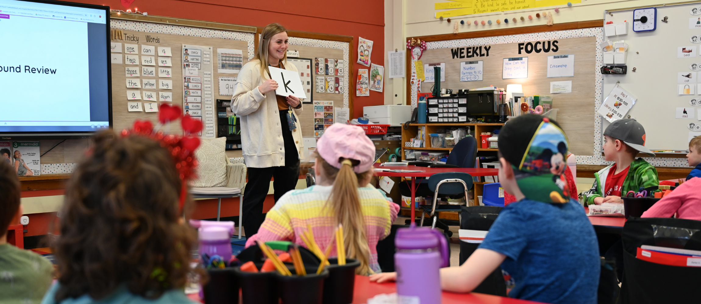 An image of a teacher at the front of a classroom holding a card with a letter in front of her students.