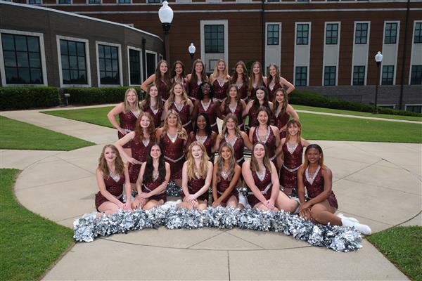 cheerleading team posing together for a group photo on their campus. They are wearing matching uniforms with pom poms.