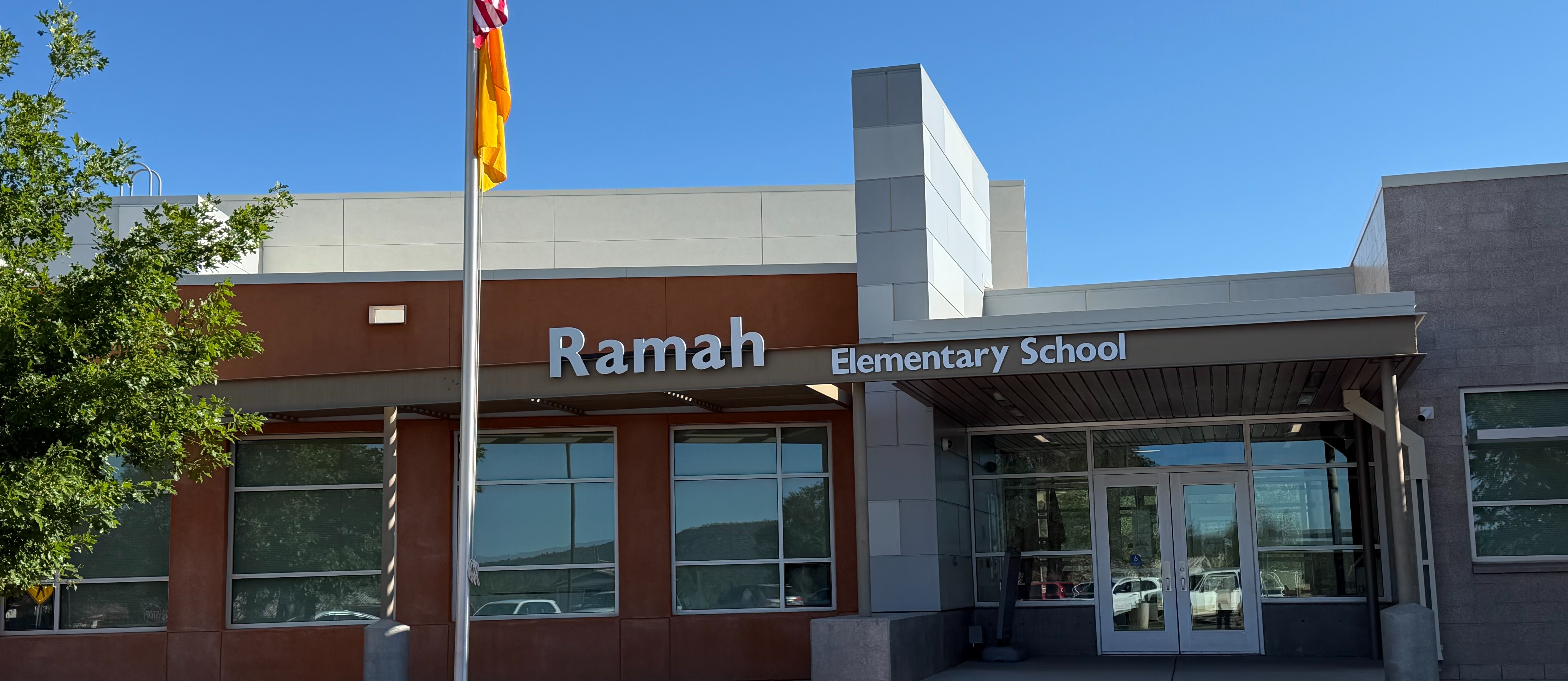 Front of Ramah Elementary School with a flag pole and sign. There are glass windows and a door.