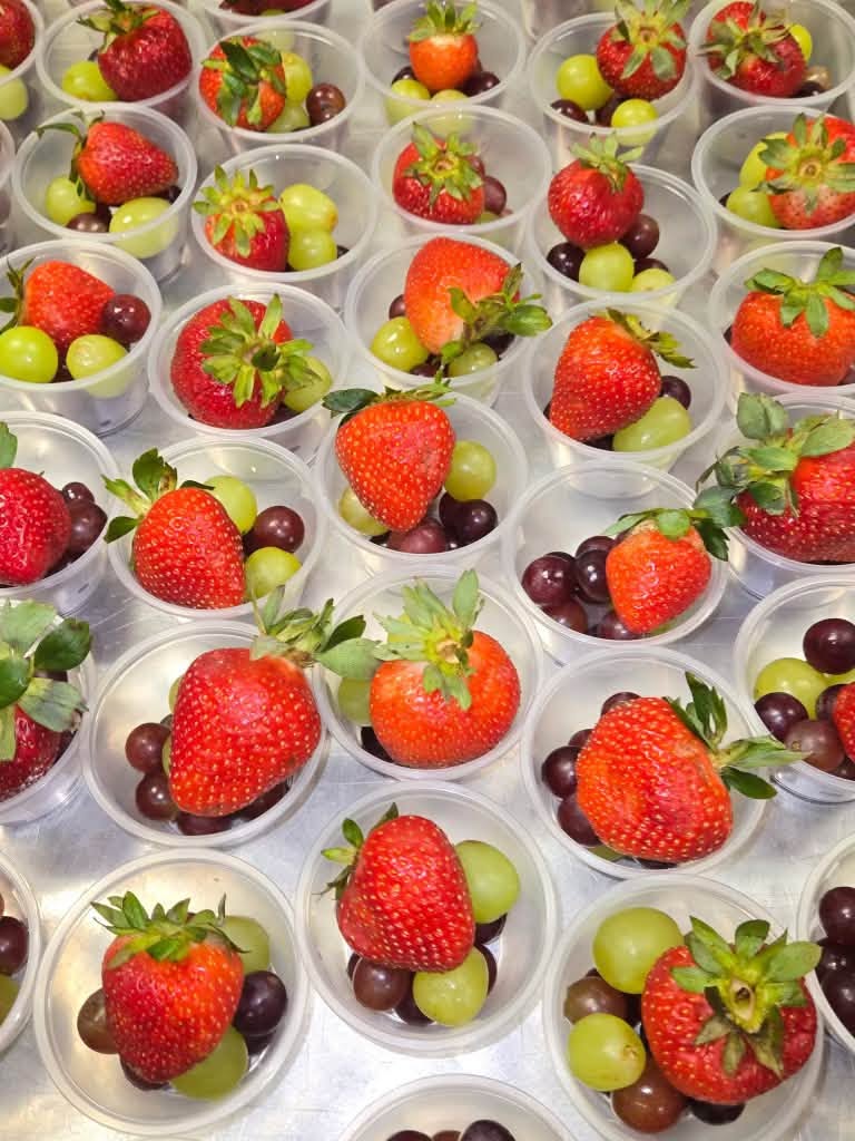 Individual cups filled with fresh strawberries and green and red grapes are arranged for student meal service.