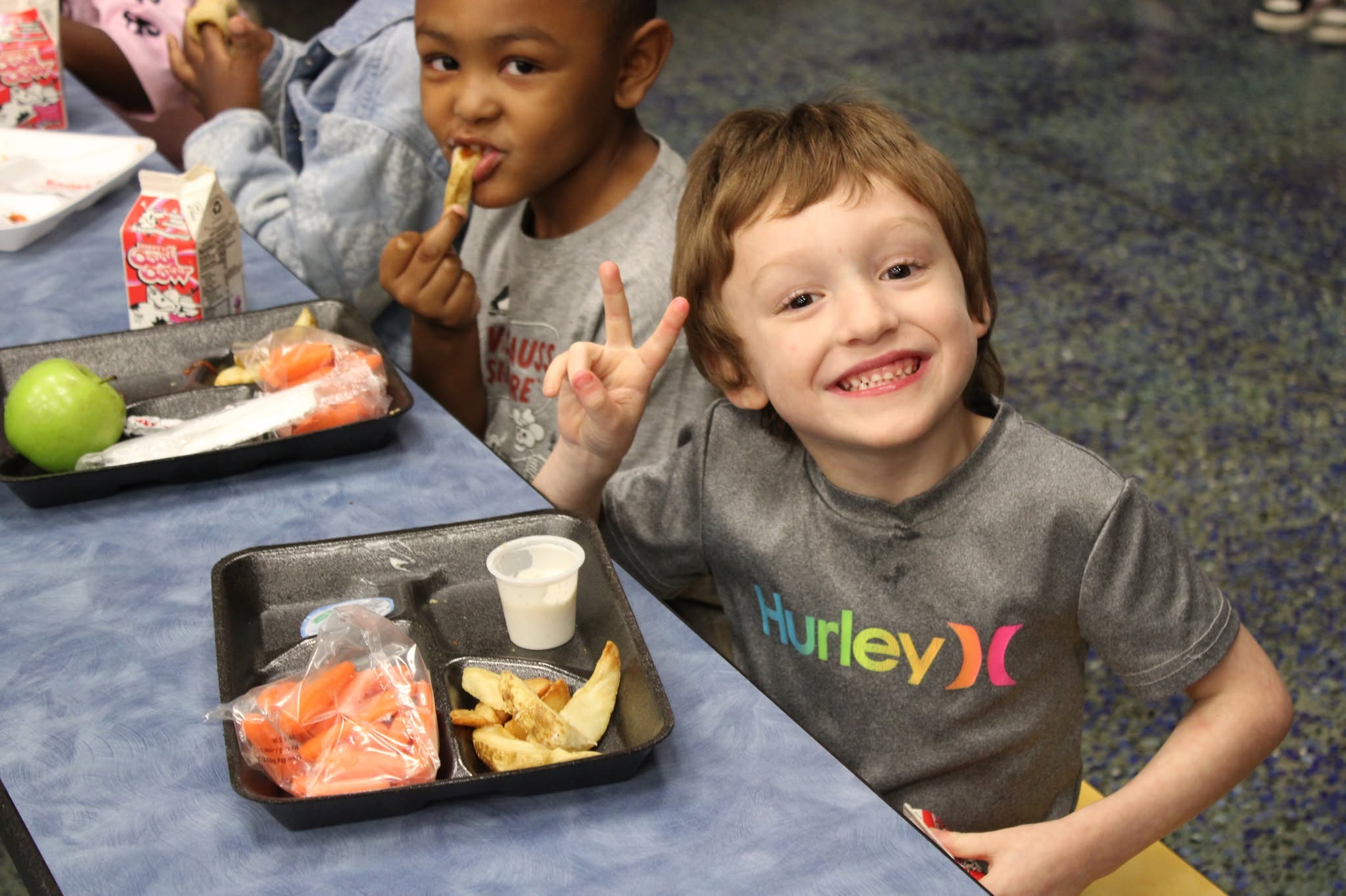 Two elementary students enjoy lunch in the cafeteria. The student in front smiles and holds up a peace sign beside a tray with potato wedges, carrots and dipping sauce.