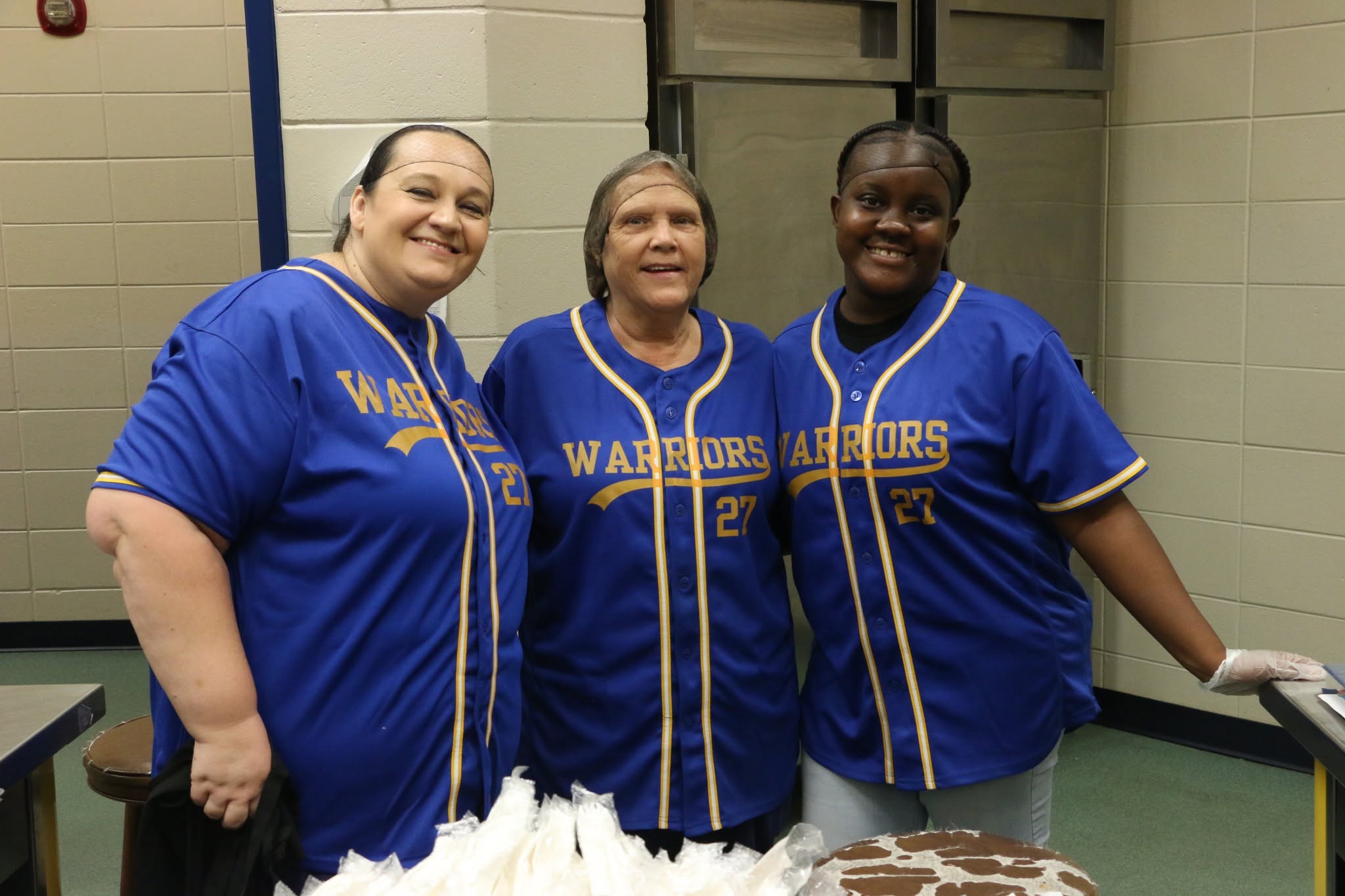 Three school nutrition team members smile together in the kitchen while wearing blue and gold Warriors jerseys.