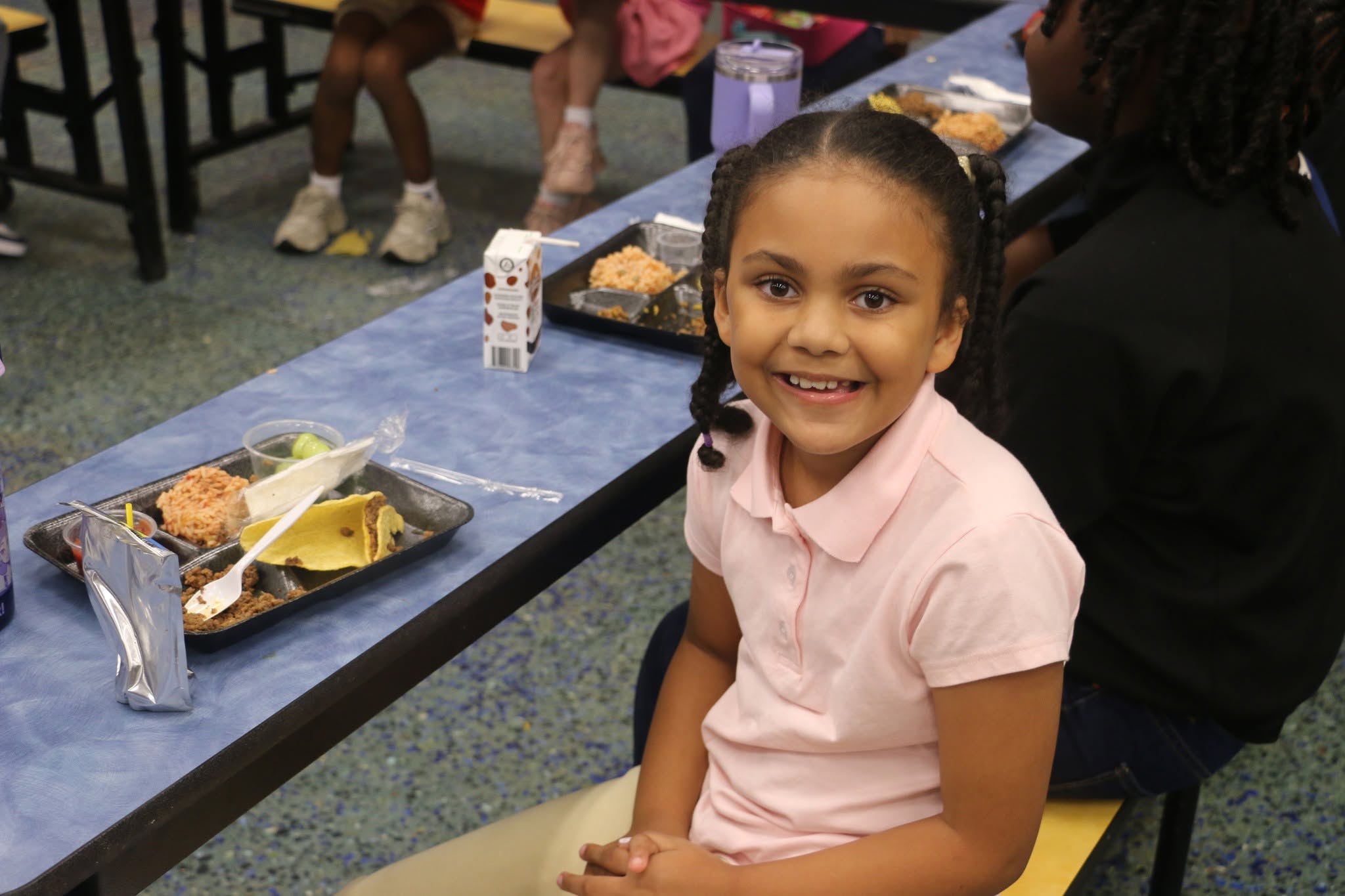 A smiling elementary student sits in the cafeteria with a lunch tray featuring tacos, rice, fruit and milk.
