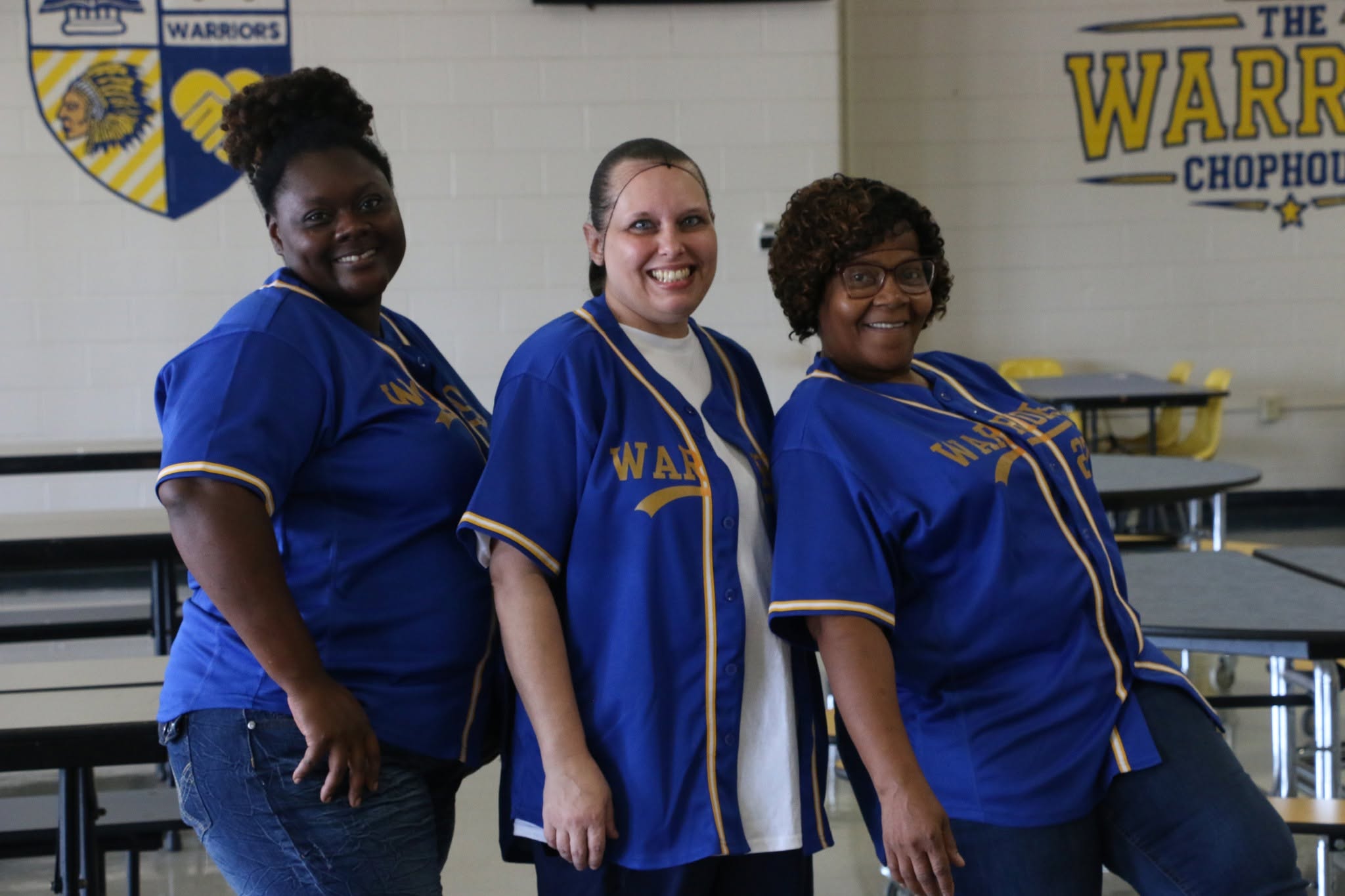 Three school nutrition team members smile together in the cafeteria while wearing blue and gold Warriors jerseys.