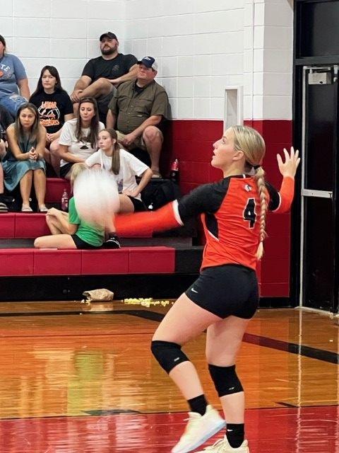 A woman in a black and orange volleyball uniform, number 4, reaching up to serve the ball.