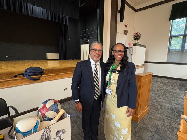 Two people stand smiling in front of a stage in an auditorium. A box with sports balls is nearby. The atmosphere is warm and professional.