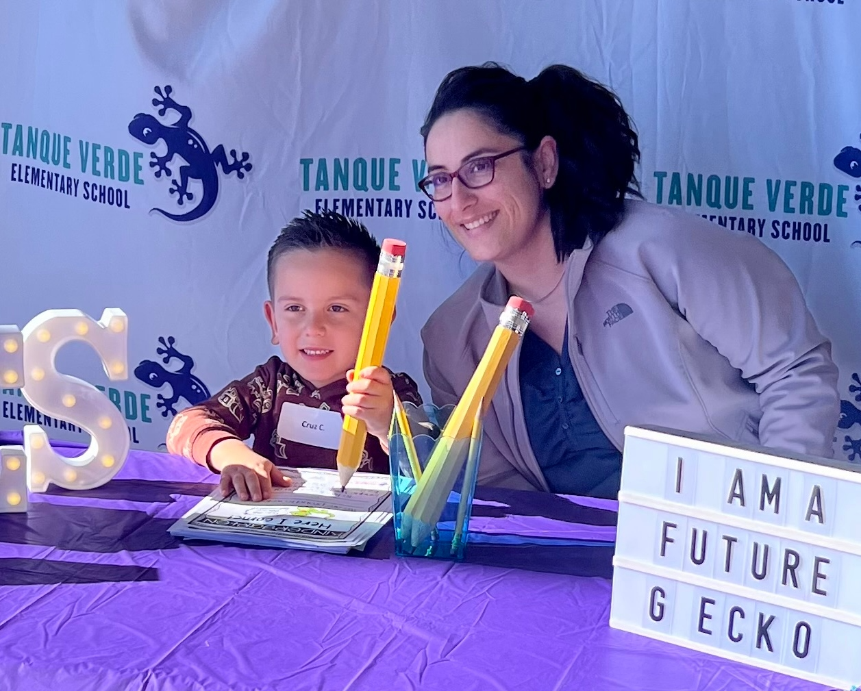 A woman and a child sit at a table with a sign reading "I am future gecko," smiling and engaged in conversation.