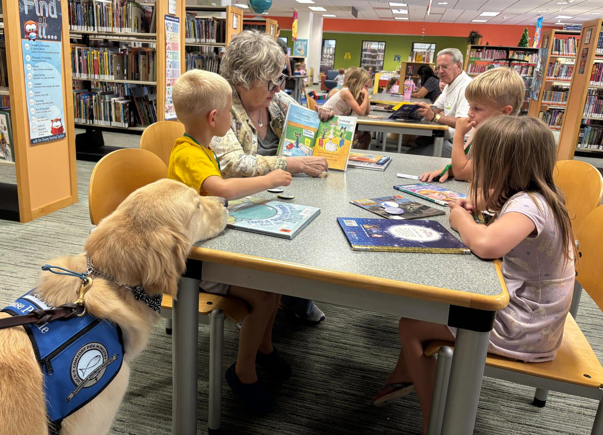 A group of kids read with a volunteer as Meshach the Comfort Dog looks on.