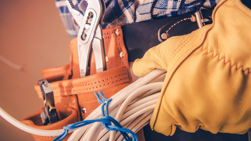 A close-up of a tradesperson's tool belt with tools and coiled wires, wearing protective gloves.