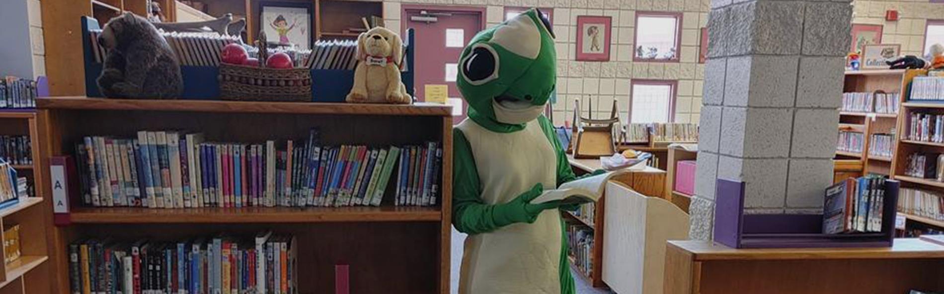 A green lizard perched on a stack of books in a library, appearing to hold a book with its front legs.