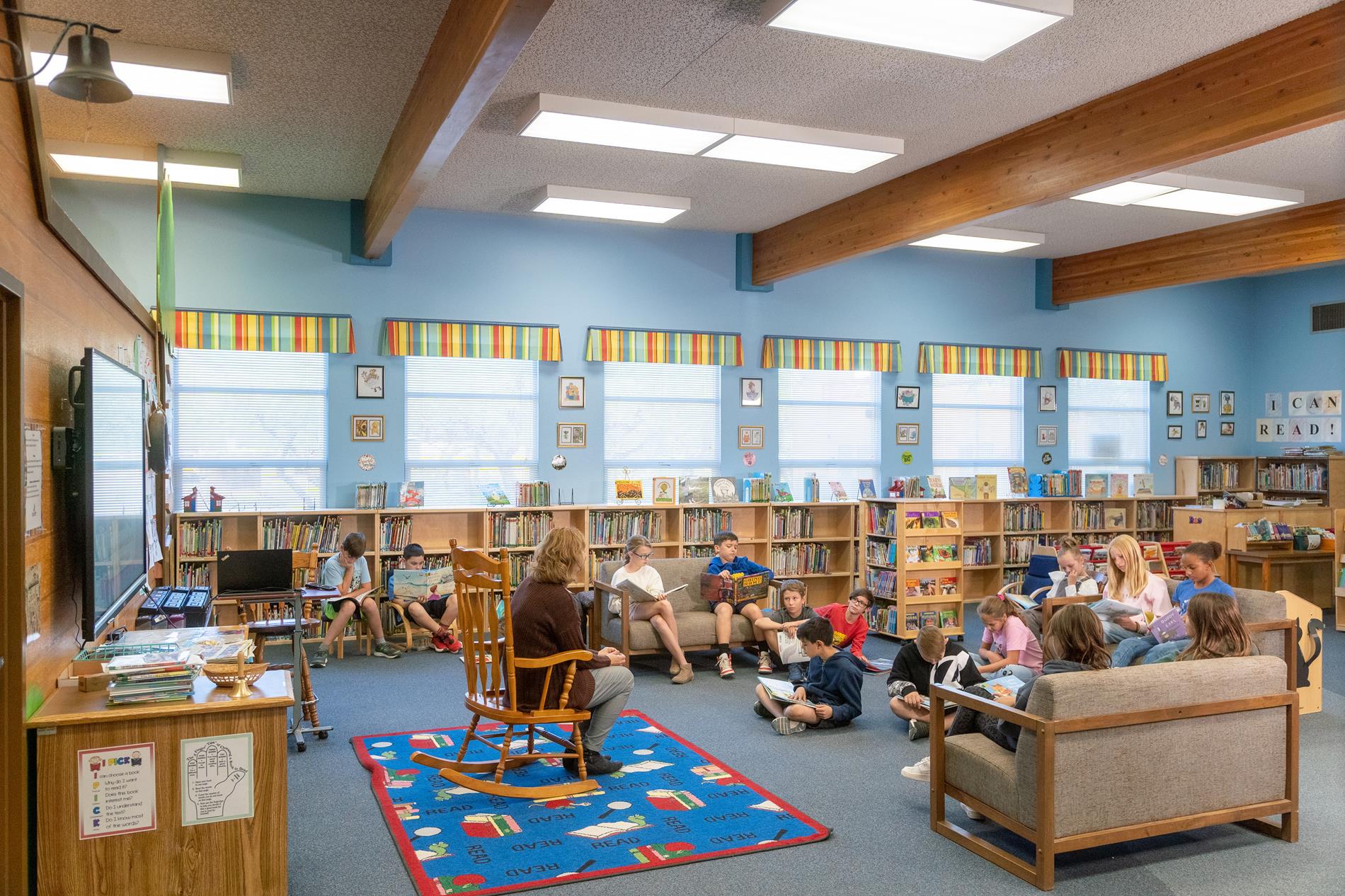 A teacher sits with a group of children in a library, engaged in reading and discussion among bookshelves.