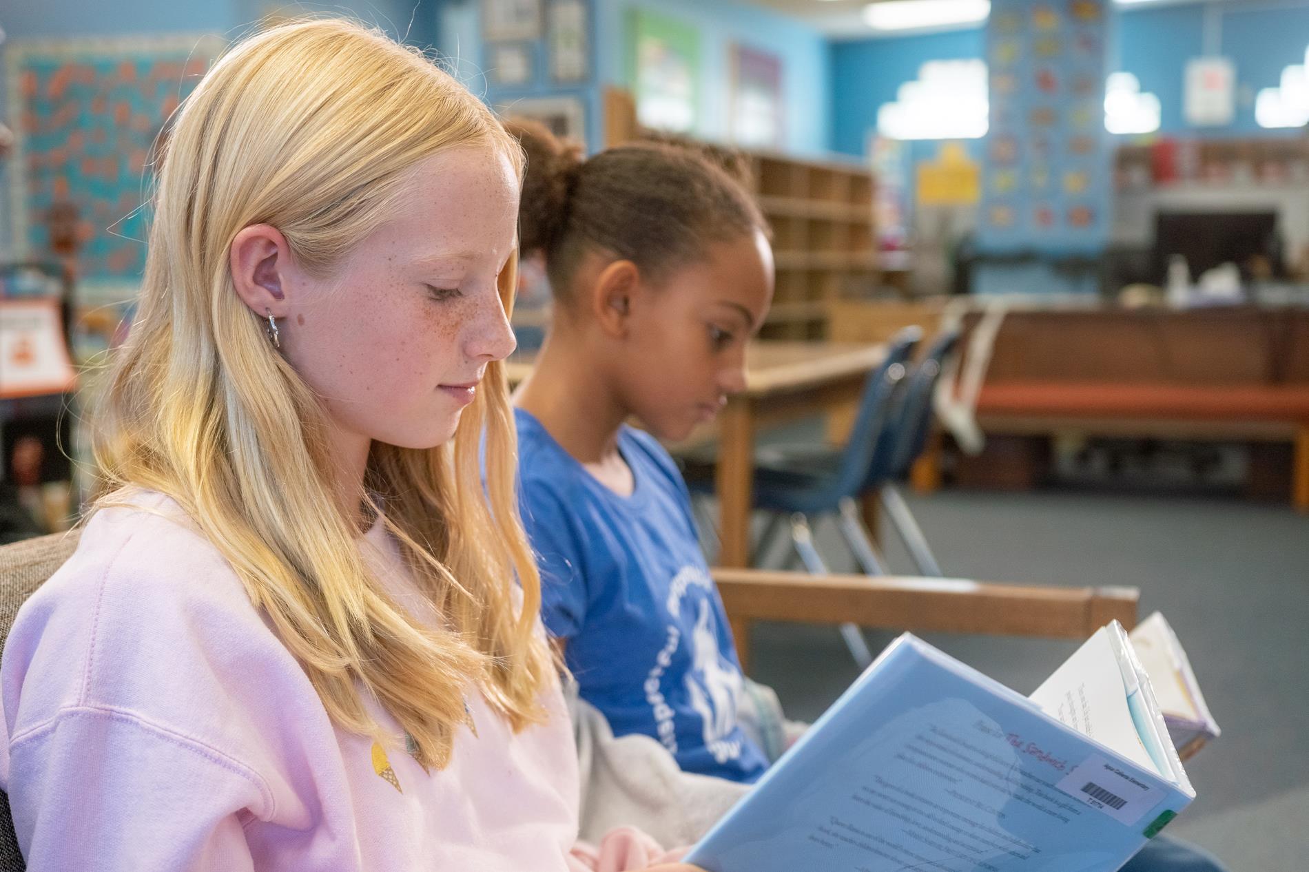 Two girls sit at a classroom desk, focused on reading books, surrounded by educational materials and a bright learning environment.
