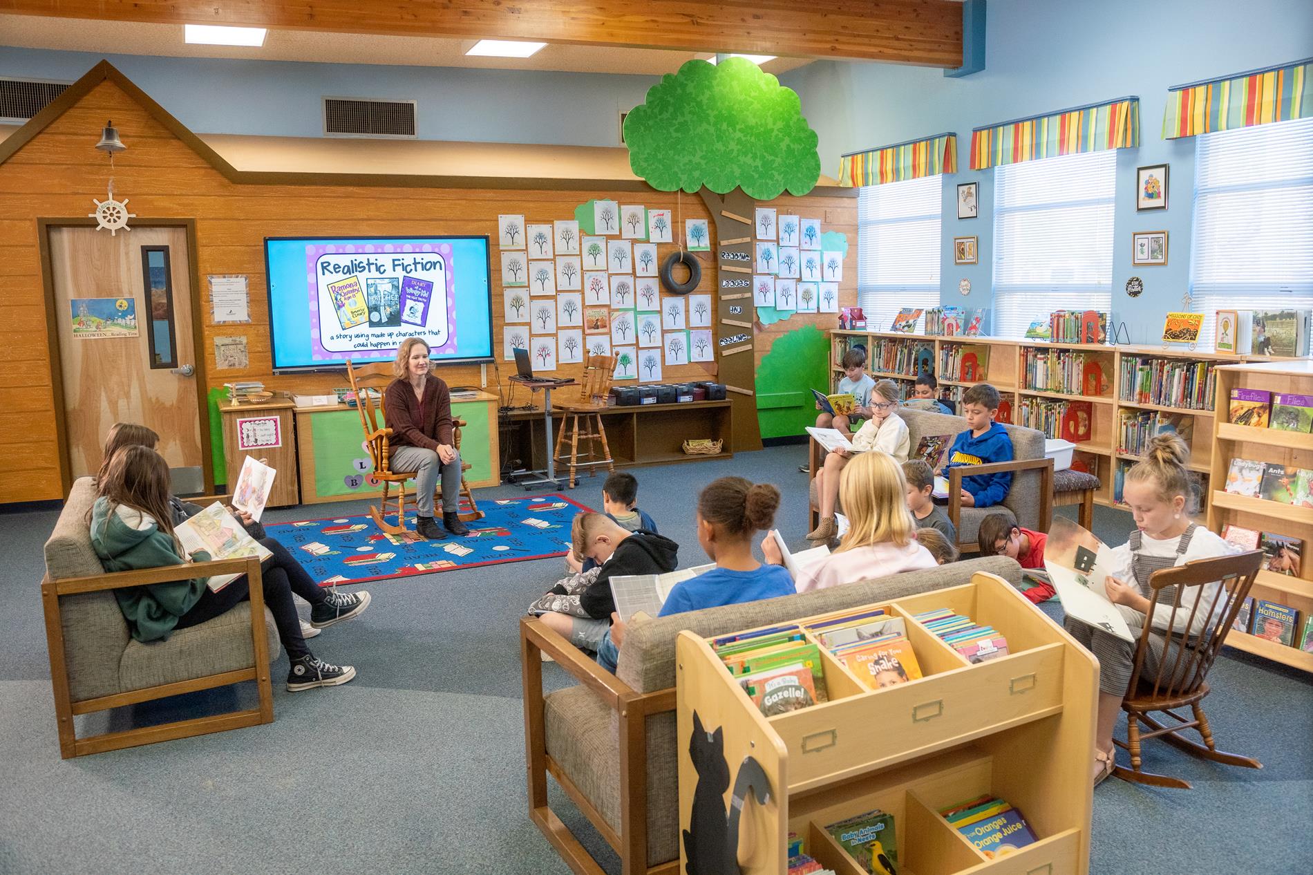 Children seated in chairs in a library, surrounded by shelves filled with various books.